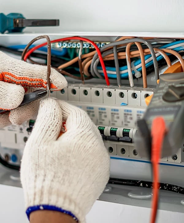 Close-up view of an electrician's hands in protective safety gloves testing connections inside an open electrical service panel with colorful wiring.