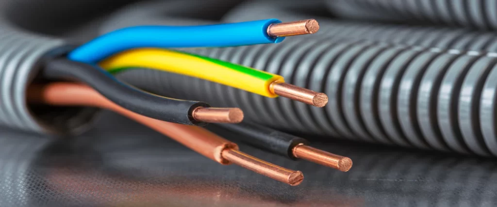 Close-up of blue, yellow, black, and brown copper electrical wires emerging from a grey corrugated flexible conduit pipe on a reflective surface.