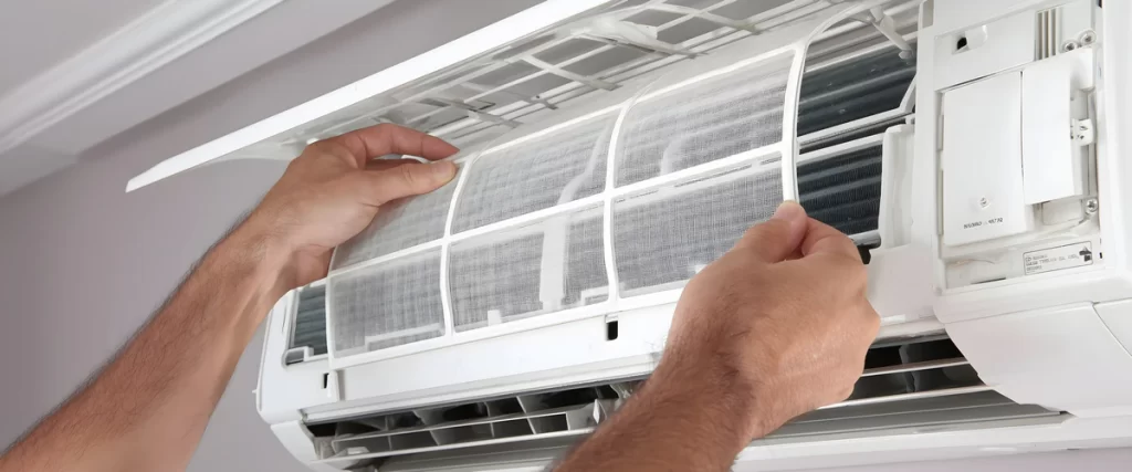 A person’s hands removing a white mesh air filter from a wall-mounted AC unit for routine maintenance and cleaning.