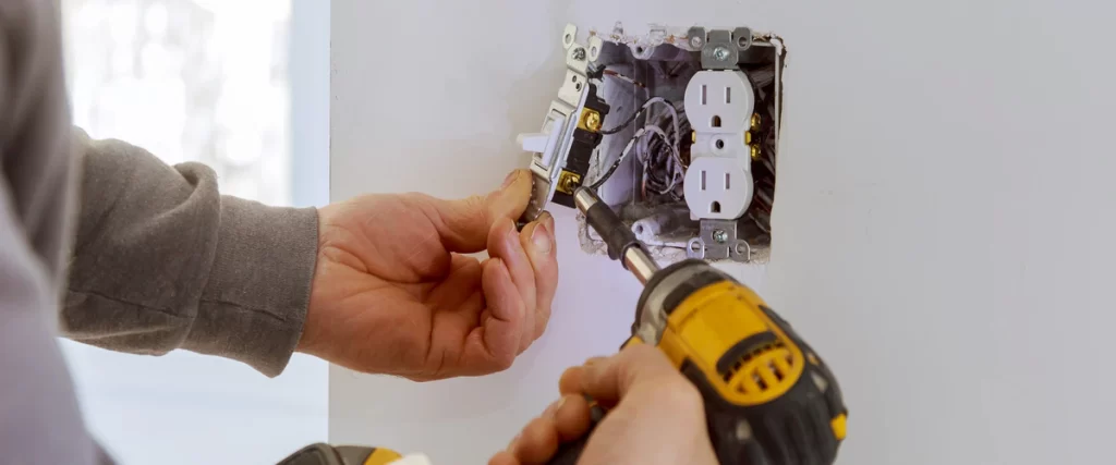 A professional electrician using a power drill to install a white light switch and double electrical outlet into a wall.